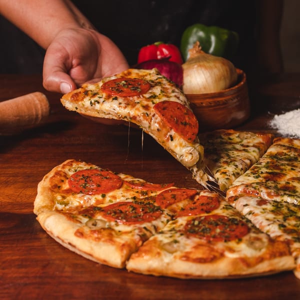 Hand lifting a cheesy pepperoni pizza slice from a whole pizza on a wooden table.