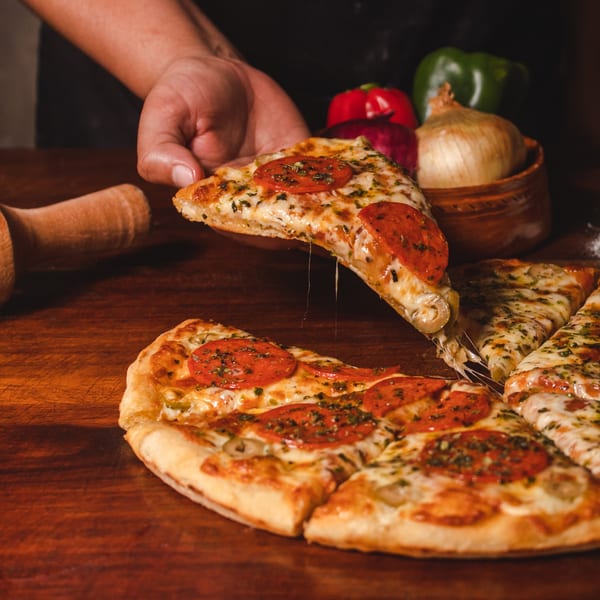 Hand lifting a cheesy pepperoni pizza slice from a whole pizza on a wooden table.